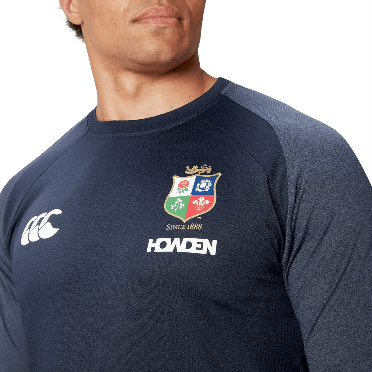 A person is wearing a dark blue sports jersey from Canterbury, featuring various logos such as a shield crest and the word "Howden", reminiscent of classic designs similar to the British &amp; Irish Lions 25 Everest Tee.