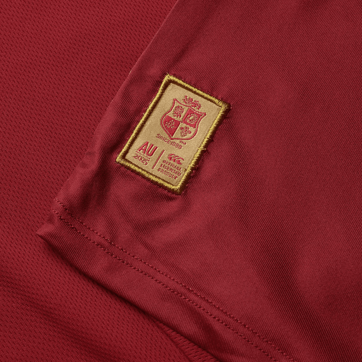 The British &amp; Irish Lions 25 Superlight Training Tee by Canterbury is a maroon sports jersey featuring a gold embroidered patch with a shield, &quot;AU 2023&quot; text, and brand logos. This tee is ideal for intense sessions and embodies Canterbury&#39;s renowned quality.
