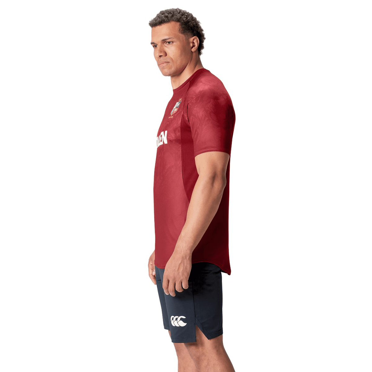 A man stands in a red British &amp; Irish Lions 25 Superlight Training Tee by Canterbury, paired with navy shorts, embodying the iconic style of the British &amp; Irish Lions with a neutral expression.