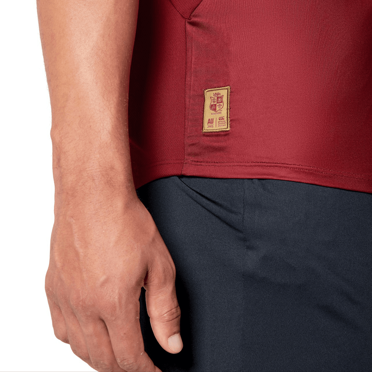 Close-up of a person wearing a red British &amp; Irish Lions 25 Superlight Training Tee by Canterbury paired with dark pants. The tee includes a beige patch featuring a crest reminiscent of the British &amp; Irish Lions and small text near the hem. The person's left arm rests calmly by their side.