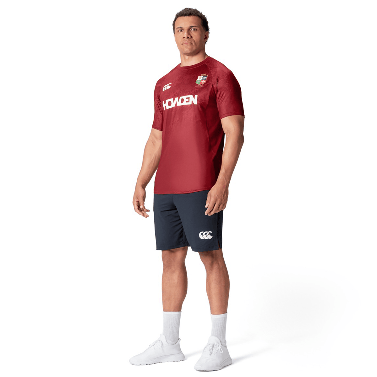 A person stands wearing a British &amp; Irish Lions 25 Superlight Training Tee by Canterbury, featuring &quot;HOWDEN&quot; across the chest, paired with navy shorts, white socks, and white sneakers.