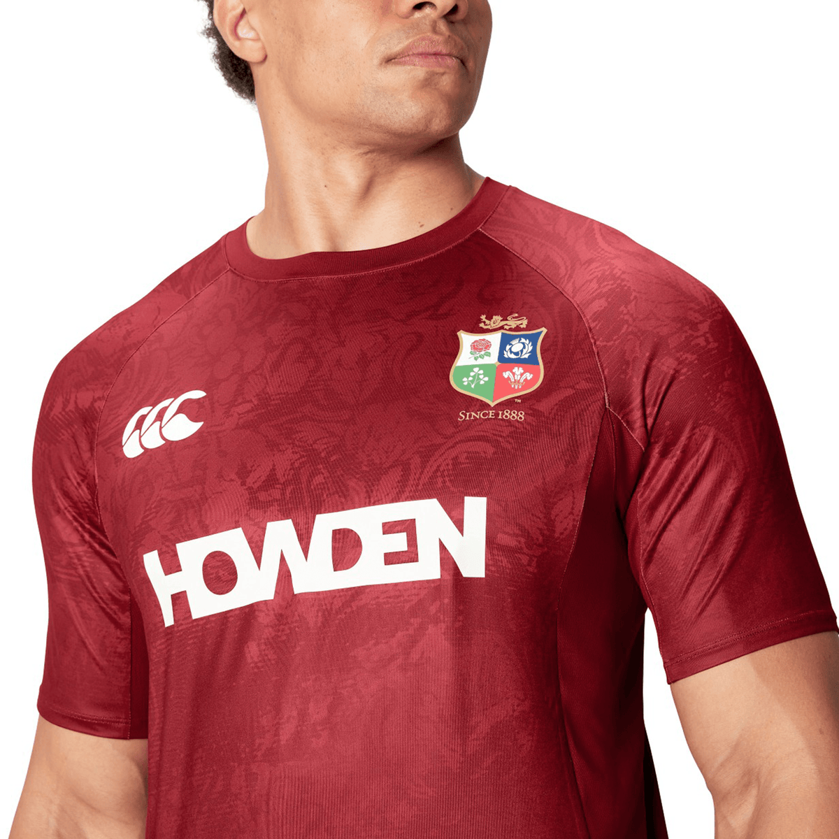 A man wears a red rugby jersey prominently displaying the &quot;HOWDEN&quot; name and team logo, reminiscent of the British &amp; Irish Lions&#39; style. This design is inspired by the British &amp; Irish Lions 25 Superlight Training Tee by Canterbury, offering fans a perfect blend of comfort and heritage.
