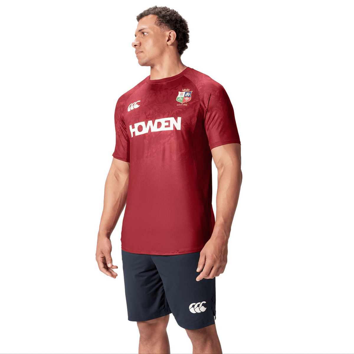 A man wearing a red British &amp; Irish Lions 25 Superlight Training Tee by Canterbury, featuring the word &quot;Howden,&quot; stands against a white background.