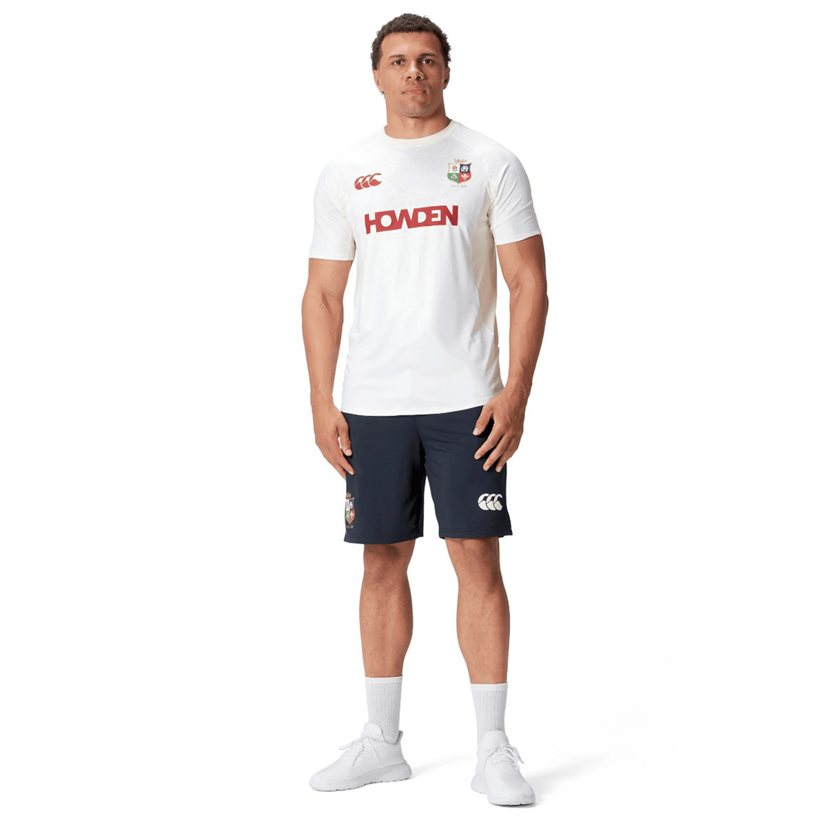 A person wearing a British &amp; Irish Lions 25 Superlight Training Tee by Canterbury, featuring a red &quot;HOWDEN&quot; logo, paired with navy shorts, white socks, and white athletic shoes stands against a plain white background.