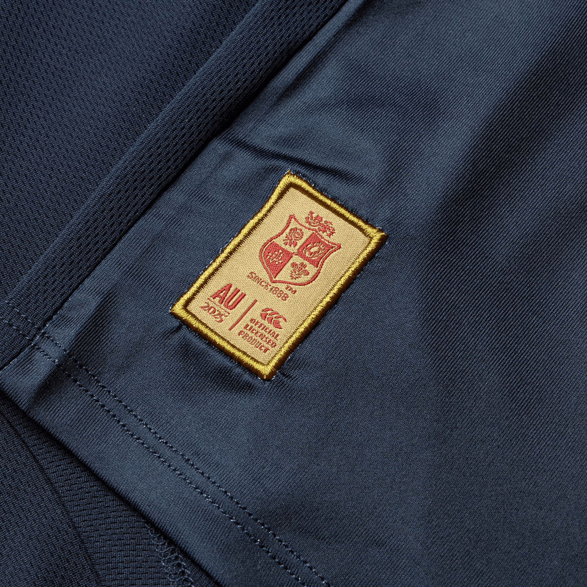 Close-up of a dark blue fabric on the British &amp; Irish Lions 25 Superlight Training Tee by Canterbury, featuring a rectangular gold patch with a red shield, &quot;since 1888,&quot; &quot;AU 2025,&quot; and other text. The patch boasts a meticulously sewn border.