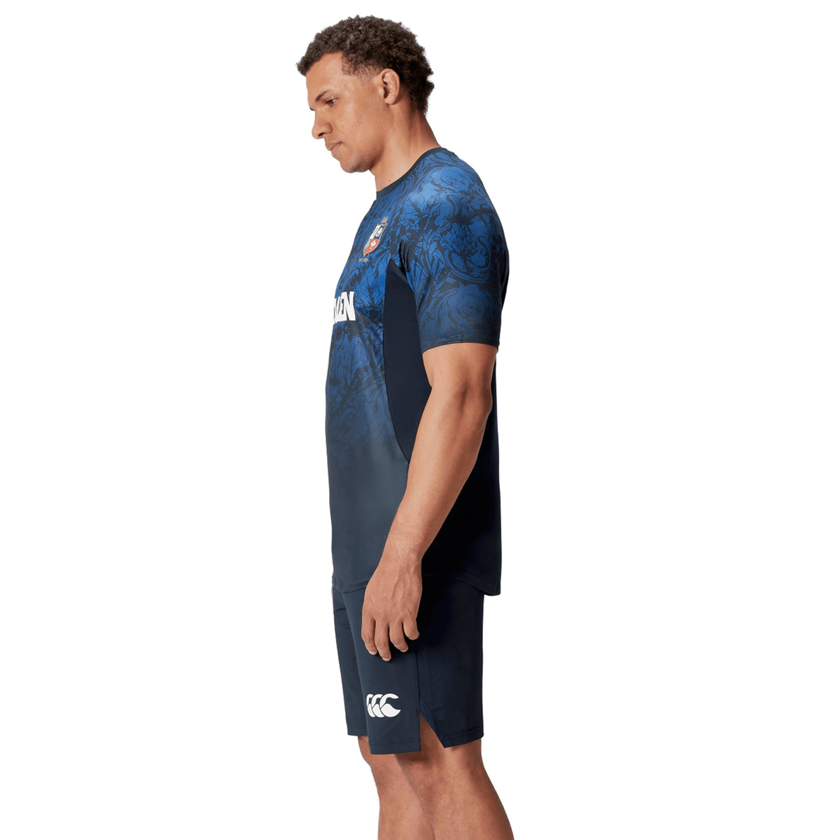 A person wearing a British &amp; Irish Lions 25 Superlight Training Tee by Canterbury, featuring a blue and black floral pattern, stands with their hands by their sides, viewed from the side.