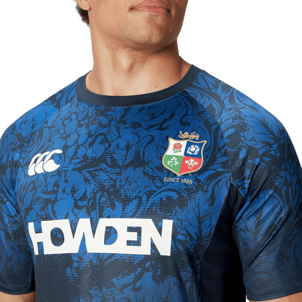 A man wearing the British &amp; Irish Lions 25 Superlight Training Tee by Canterbury captures the essence of the team with its blue design, floral patterns, team crest, and various logos.
