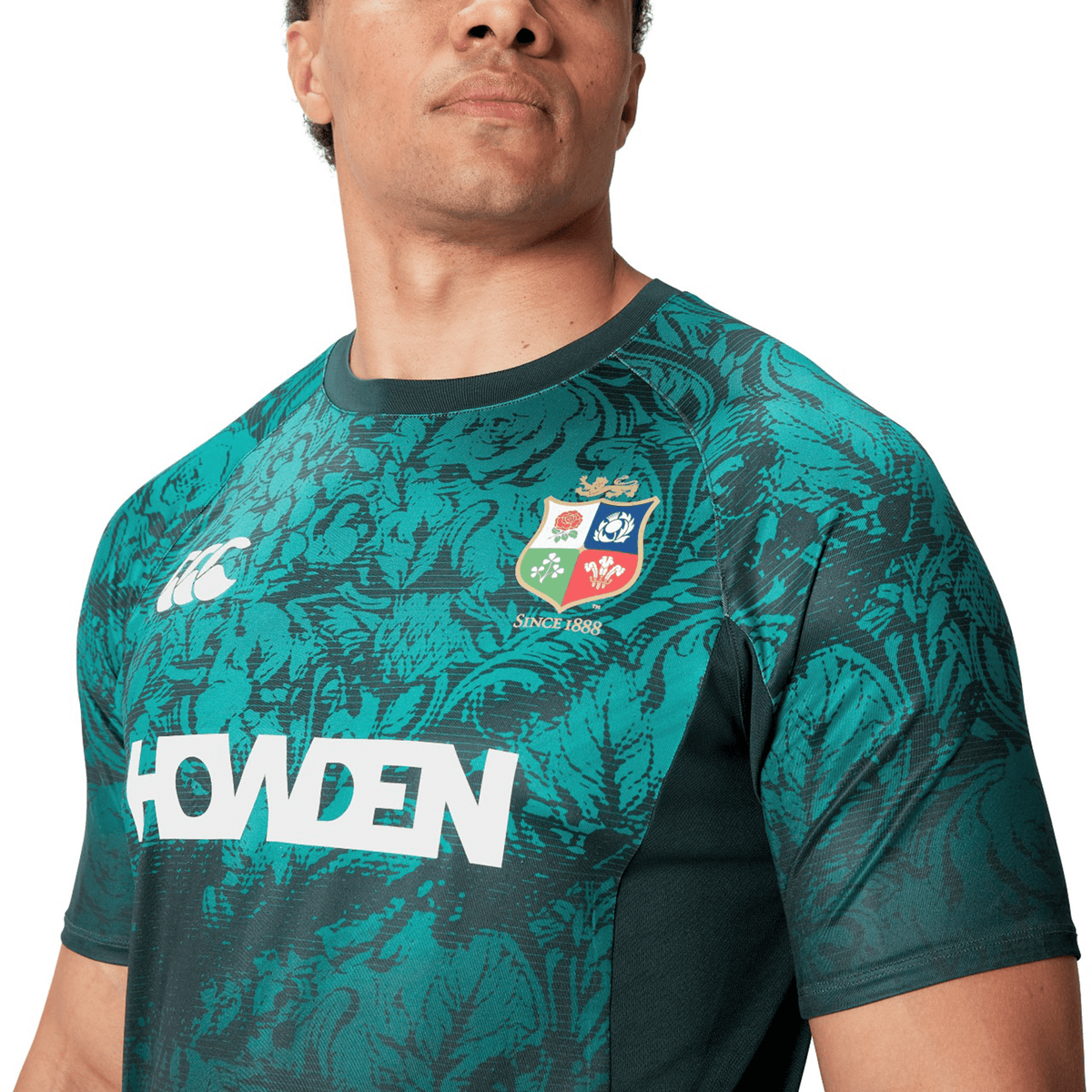 A person wearing the British &amp; Irish Lions 25 Superlight Training Tee by Canterbury, featuring a green rugby jersey with a patterned design and logos, including &quot;Howden&quot; and a crest badge, reminiscent of the iconic style by Canterbury.