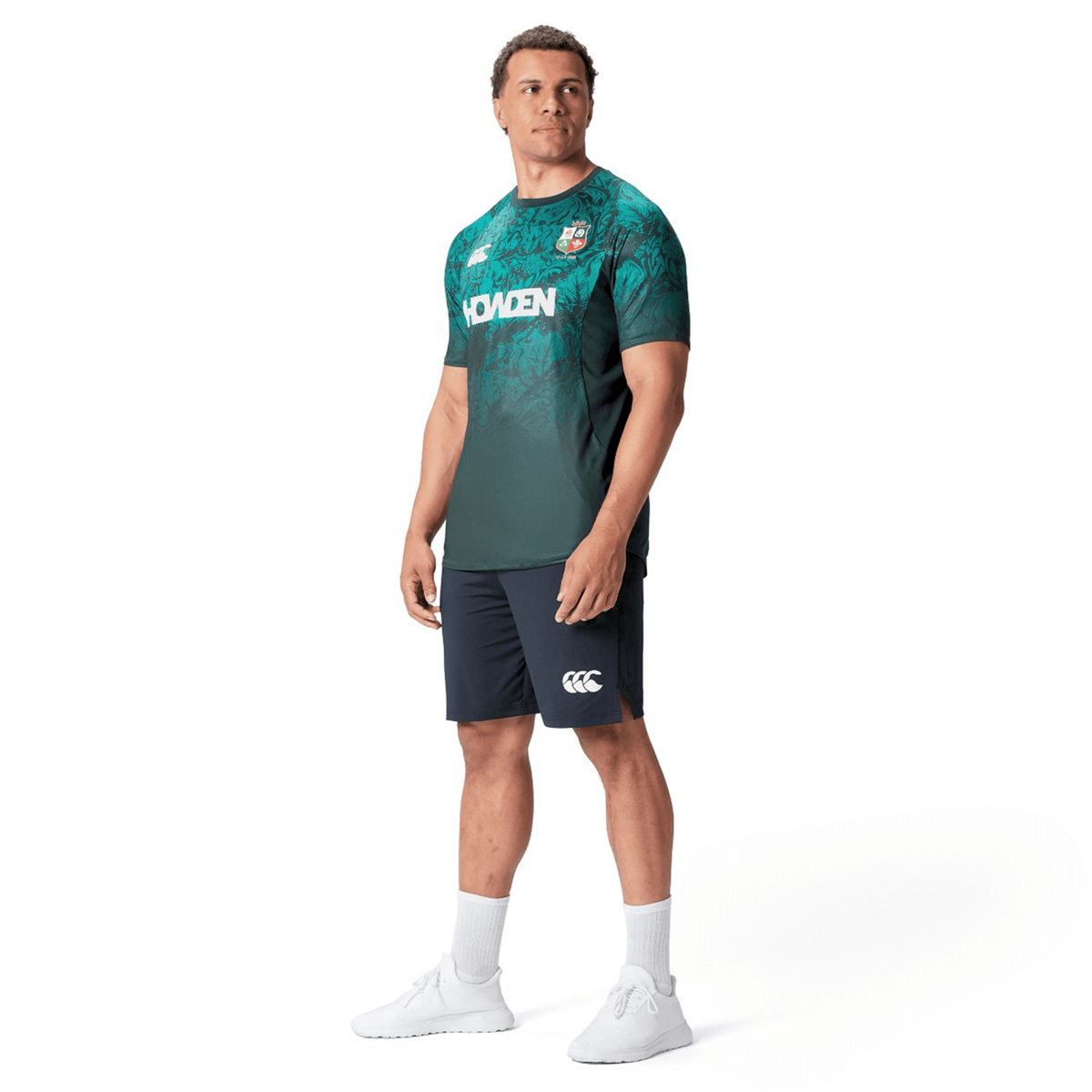 A man wearing a green and blue jersey, reminiscent of the British &amp; Irish Lions 25 Superlight Training Tee by Canterbury, and navy shorts stands facing slightly to the left. He is sporting white socks and shoes against a plain white background.