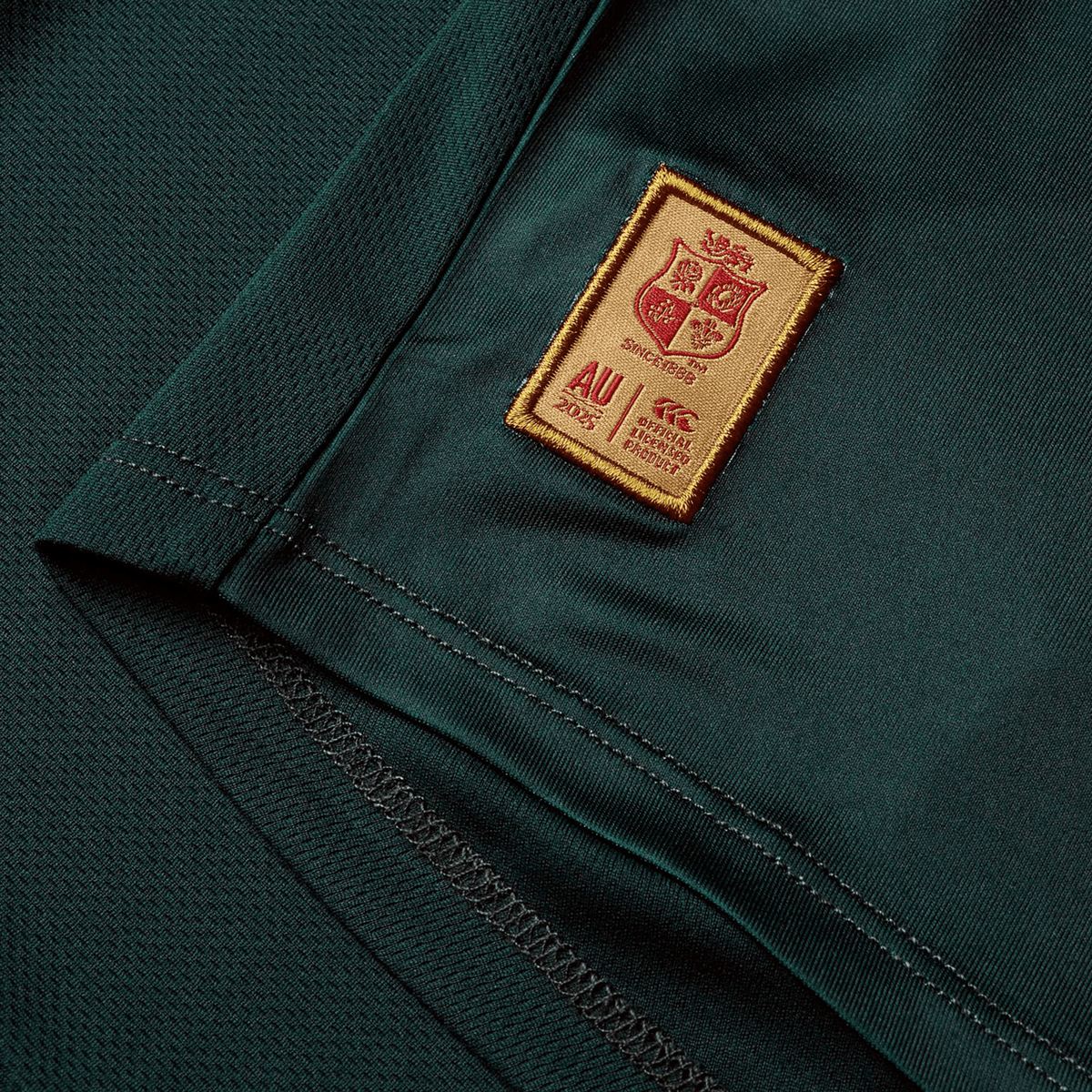 A close-up view of a deep green fabric features a sewn red and gold patch with a crest, text, and the date &quot;AU 2023.&quot; Similar to the exceptional craftsmanship of Canterbury, this fabric showcases visible stitching and a textured pattern reminiscent of the British &amp; Irish Lions 25 Superlight Training Tee by Canterbury.