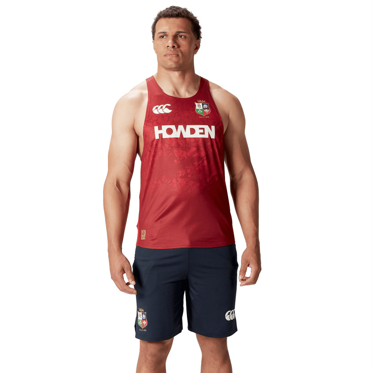 A person wears a British &amp; Irish Lions 25 Poly Singlet by Canterbury in red, paired with navy shorts, both featuring the logos.