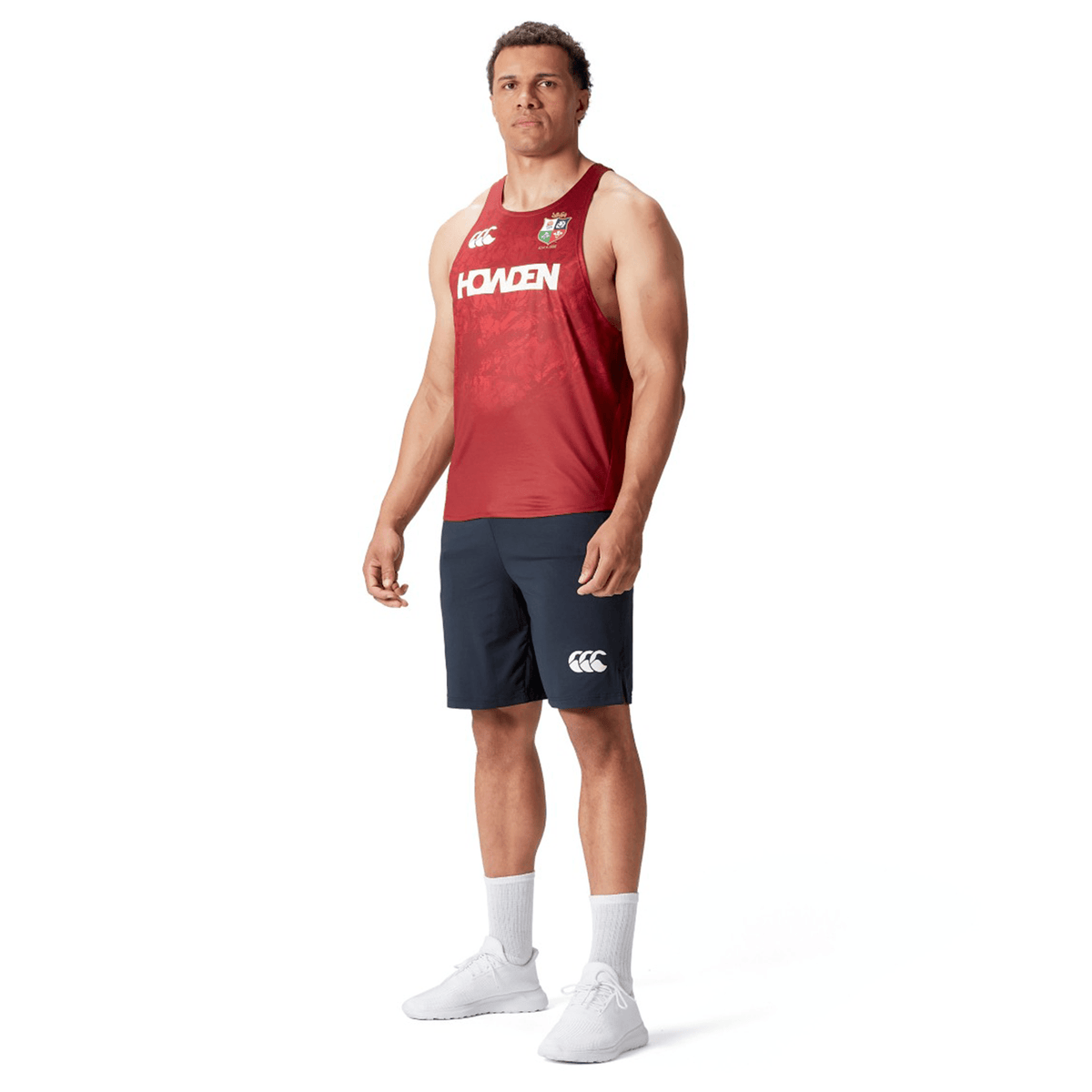 A person wearing a red British &amp; Irish Lions 25 Poly Singlet by Canterbury, navy blue shorts, white socks, and white sneakers stands against a white background.