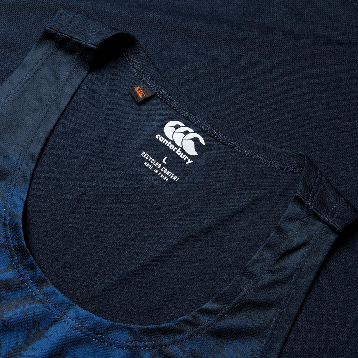 Close-up of a navy blue British &amp; Irish Lions 25 Poly Singlet by Canterbury tank top featuring a "Recycled Content" tag and size "L." The tank top rests on a similar dark blue fabric background, evoking the spirit of the British &amp; Irish Lions with its deep shade.