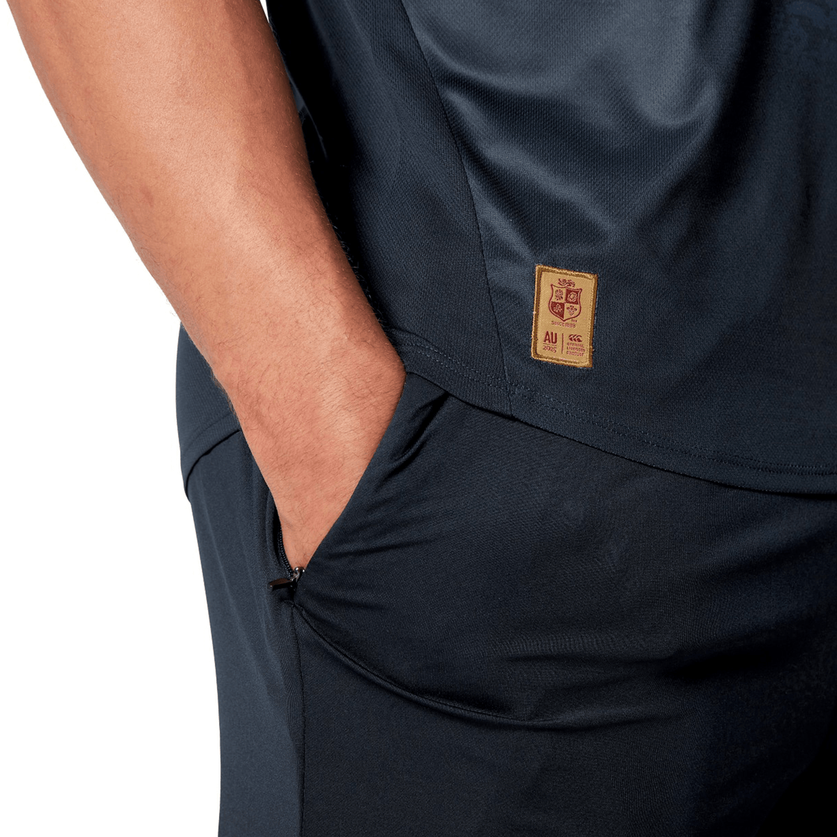 A close-up shows a person in a dark sports outfit with their hand in their pocket, wearing the stylish British &amp; Irish Lions 25 Poly Singlet by Canterbury. A small tag displaying the brand's logo and the letter "AU" is visible on the shirt, evoking the classic design of the British &amp; Irish Lions.