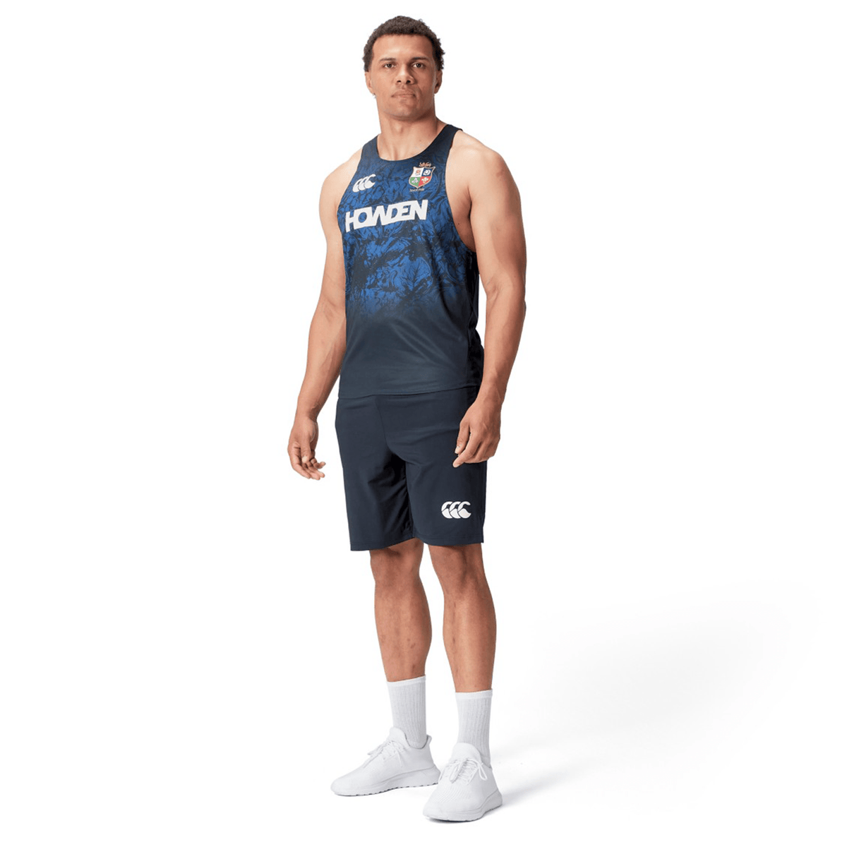 A man in the British &amp; Irish Lions 25 Poly Singlet by Canterbury sports navy athletic wear and white sneakers, standing against a white background and capturing the essence of the British &amp; Irish Lions.