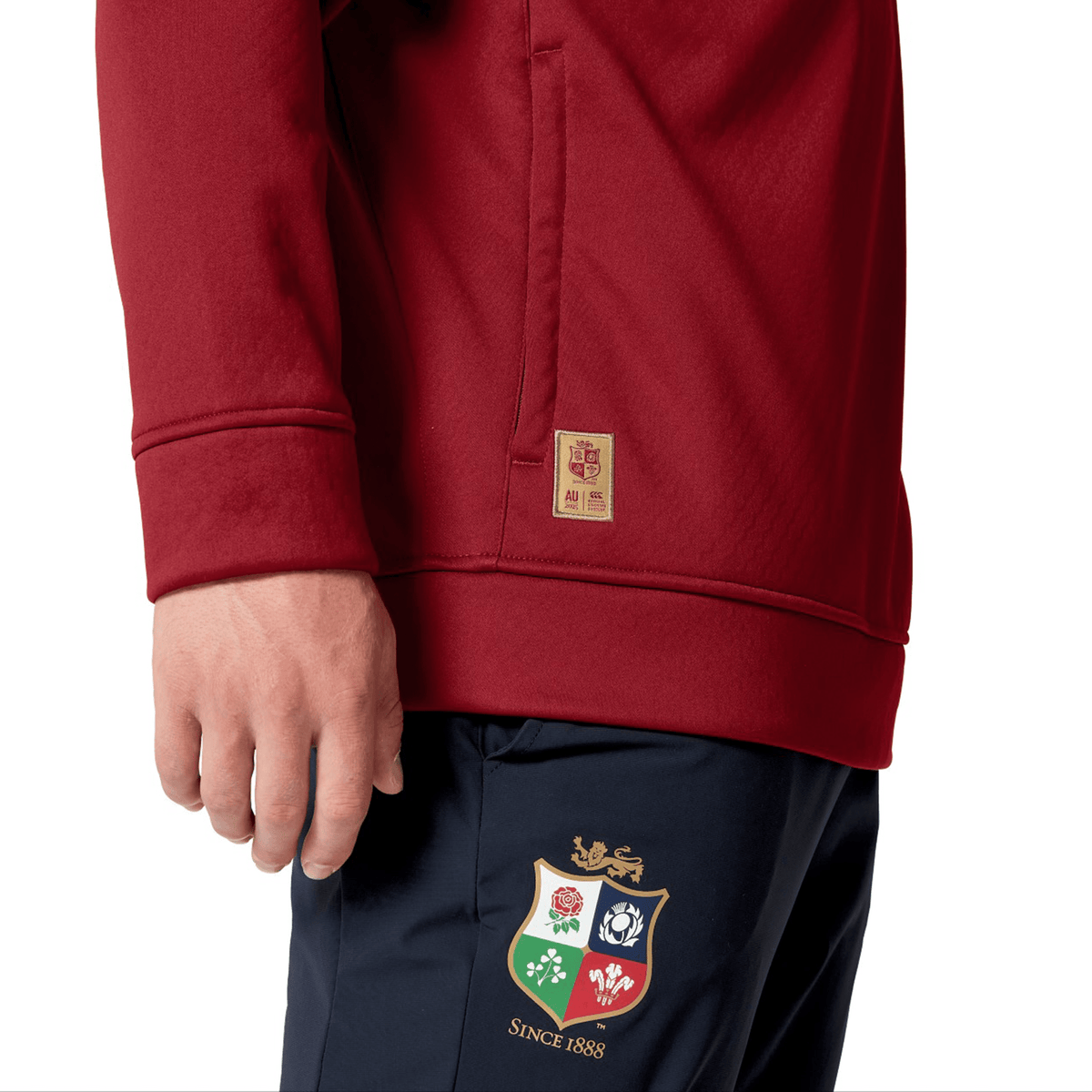 A person is wearing the British &amp; Irish Lions 2025 1/4 Zip Fleece by Canterbury, with maroon and navy colors, featuring a colorful shield emblem and &quot;Since 1888&quot; text, echoing the style of the team’s attire.