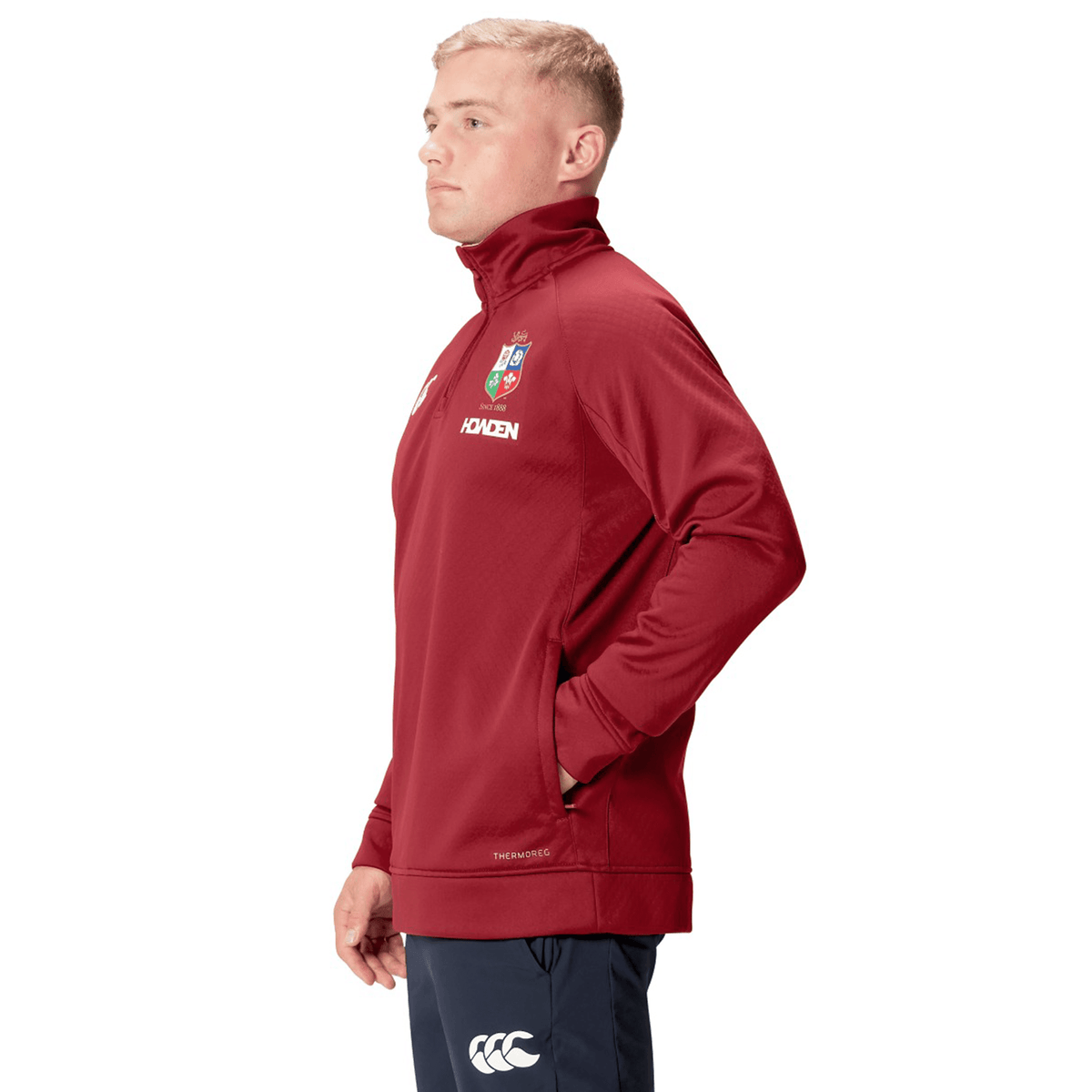 A person in the British &amp; Irish Lions 2025 1/4 Zip Fleece by Canterbury stands sideways with hands in pockets, proudly showcasing their affiliation with the British &amp; Irish Lions.