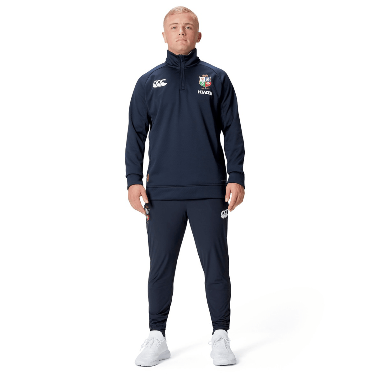 A person dressed in a navy British &amp; Irish Lions 2025 1/4 Zip Fleece by Canterbury and white shoes stands against a clean white background, exuding the timeless elegance associated with the British &amp; Irish Lions.