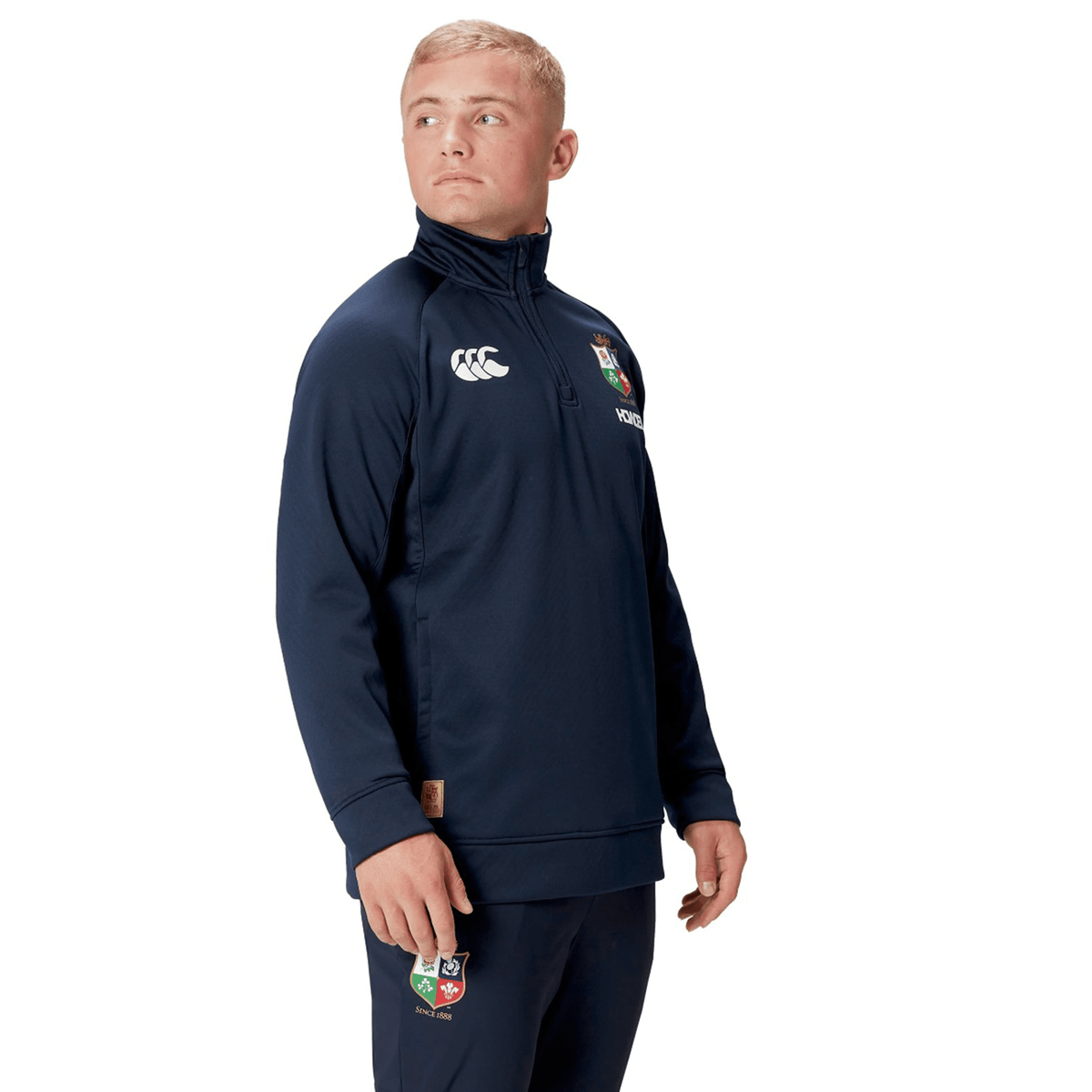 A person clad in the British &amp; Irish Lions 2025 1/4 Zip Fleece by Canterbury, featuring a logo on the chest and pants, stands against a white background.
