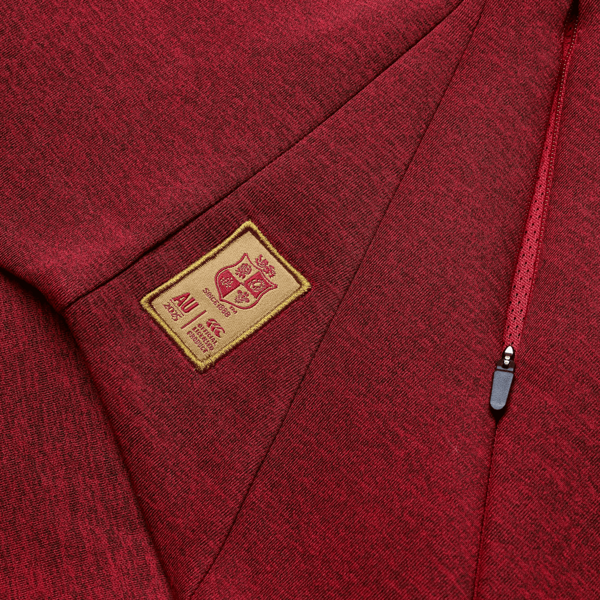 Close-up of a red fabric with a zipper, reminiscent of the British &amp; Irish Lions 2025 Everest Hoodie by Canterbury, and a beige patch featuring a crest design and text.
