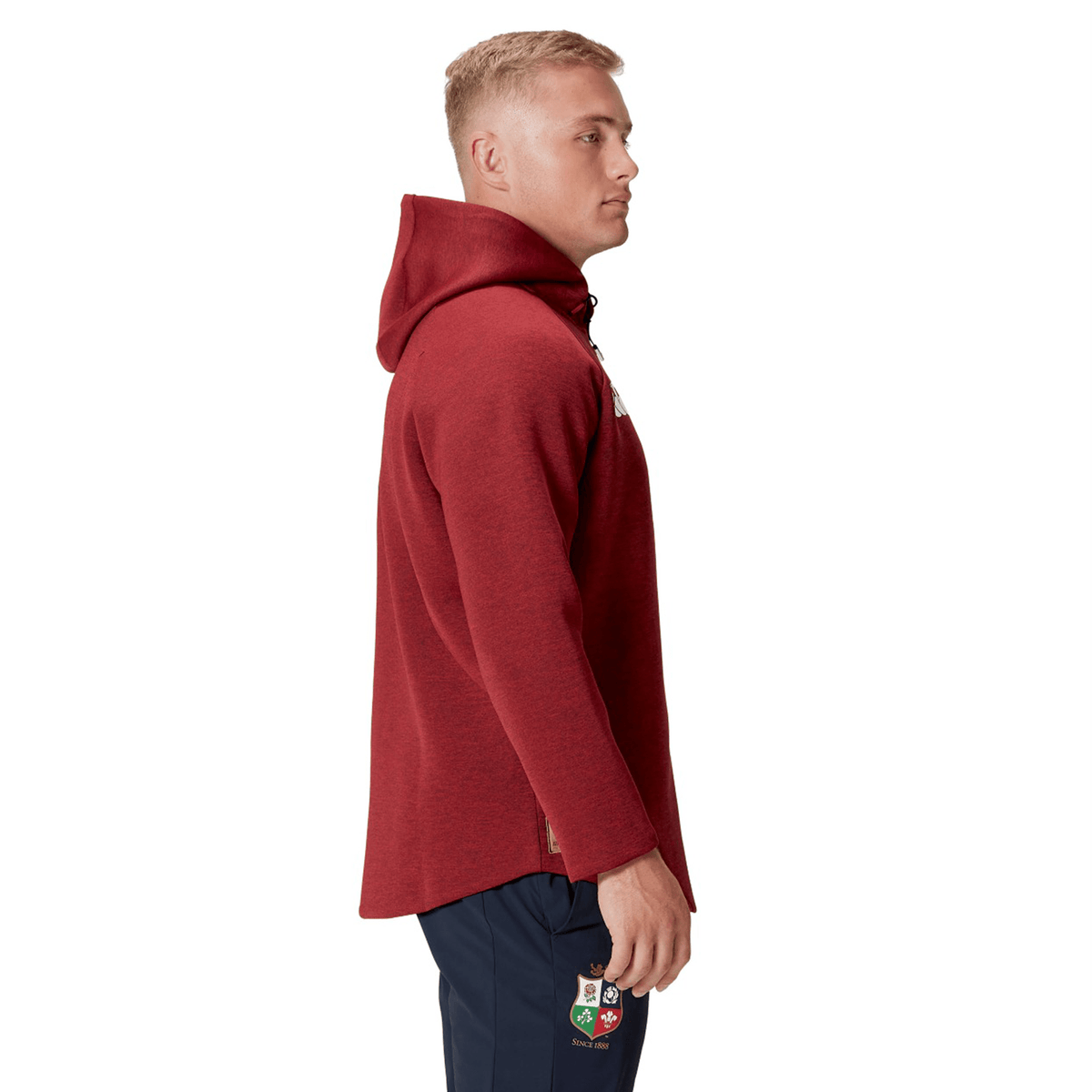 A person wearing a British &amp; Irish Lions 2025 Everest Hoodie by Canterbury and dark pants stands sideways, highlighting the logo on the pants.