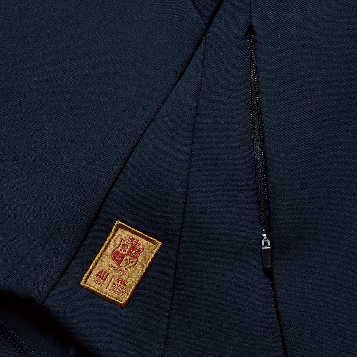 Close-up of a navy blue jacket crafted from recycled polyester, featuring a small patch with a coat of arms and "AU 2009" text, alongside a nearby zipper pocket reminiscent of the British &amp; Irish Lions 2025 Everest Hoodie by Canterbury style.