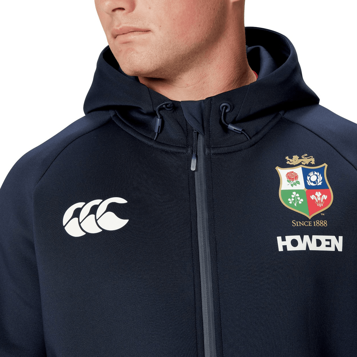 A person wearing a British &amp; Irish Lions 2025 Everest Hoodie by Canterbury, featuring logos such as "HOWDEN" and a shield emblem, crafted from recycled polyester.