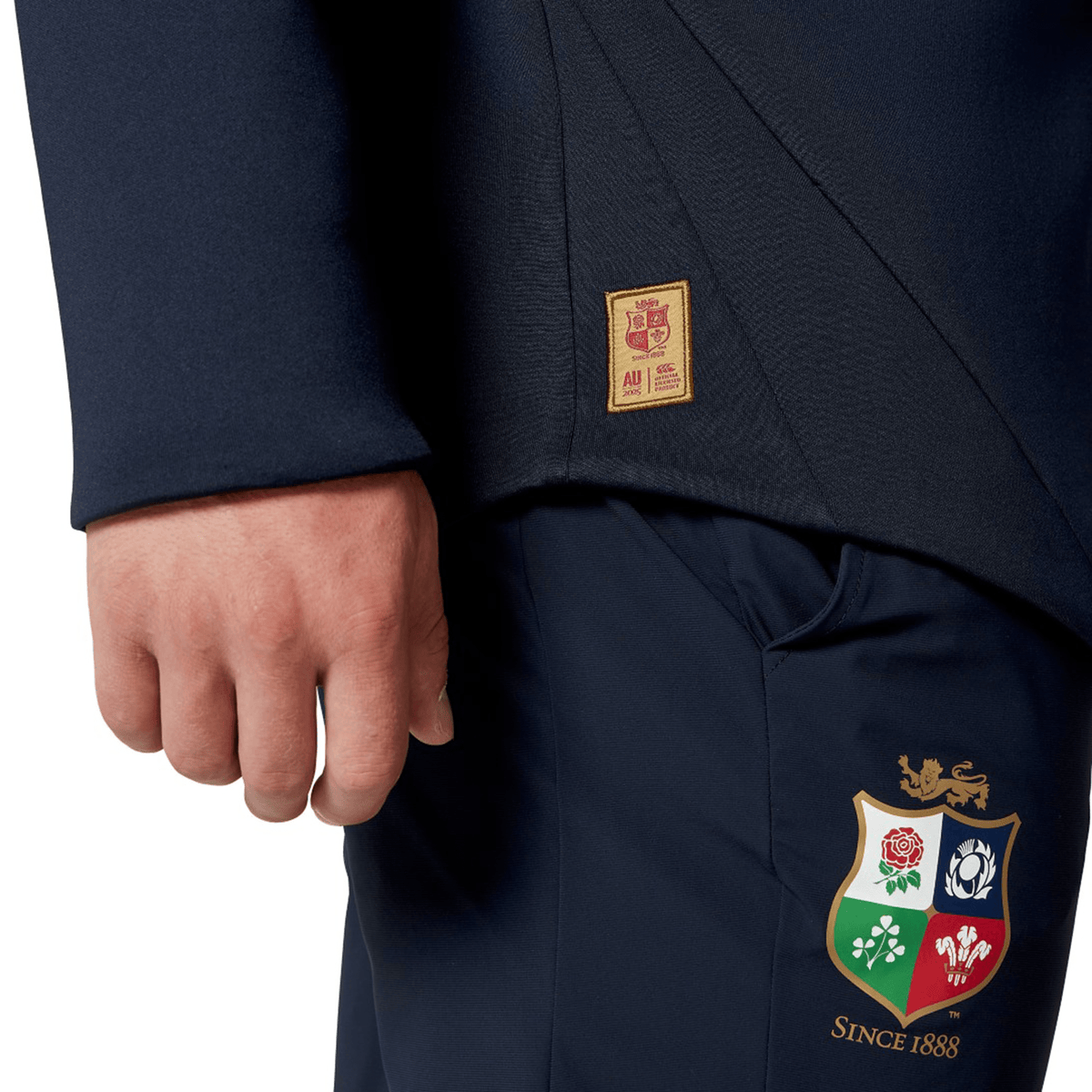 A close-up shows someone dressed in the British &amp; Irish Lions 2025 Everest Hoodie by Canterbury, designed in navy blue with recycled polyester. The pants boast the British &amp; Irish Lions logo and "Since 1888," accompanied by a small badge near the pocket.