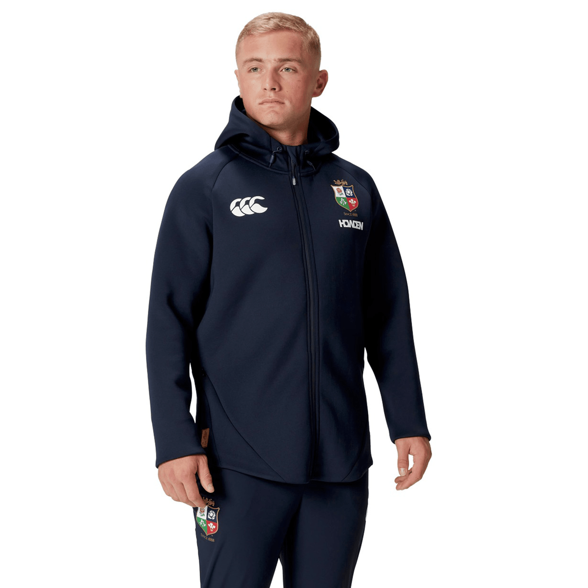 A person is wearing the British &amp; Irish Lions 2025 Everest Hoodie by Canterbury, featuring a dark blue design with embroidered logos and crafted from recycled polyester, standing against a plain white background.