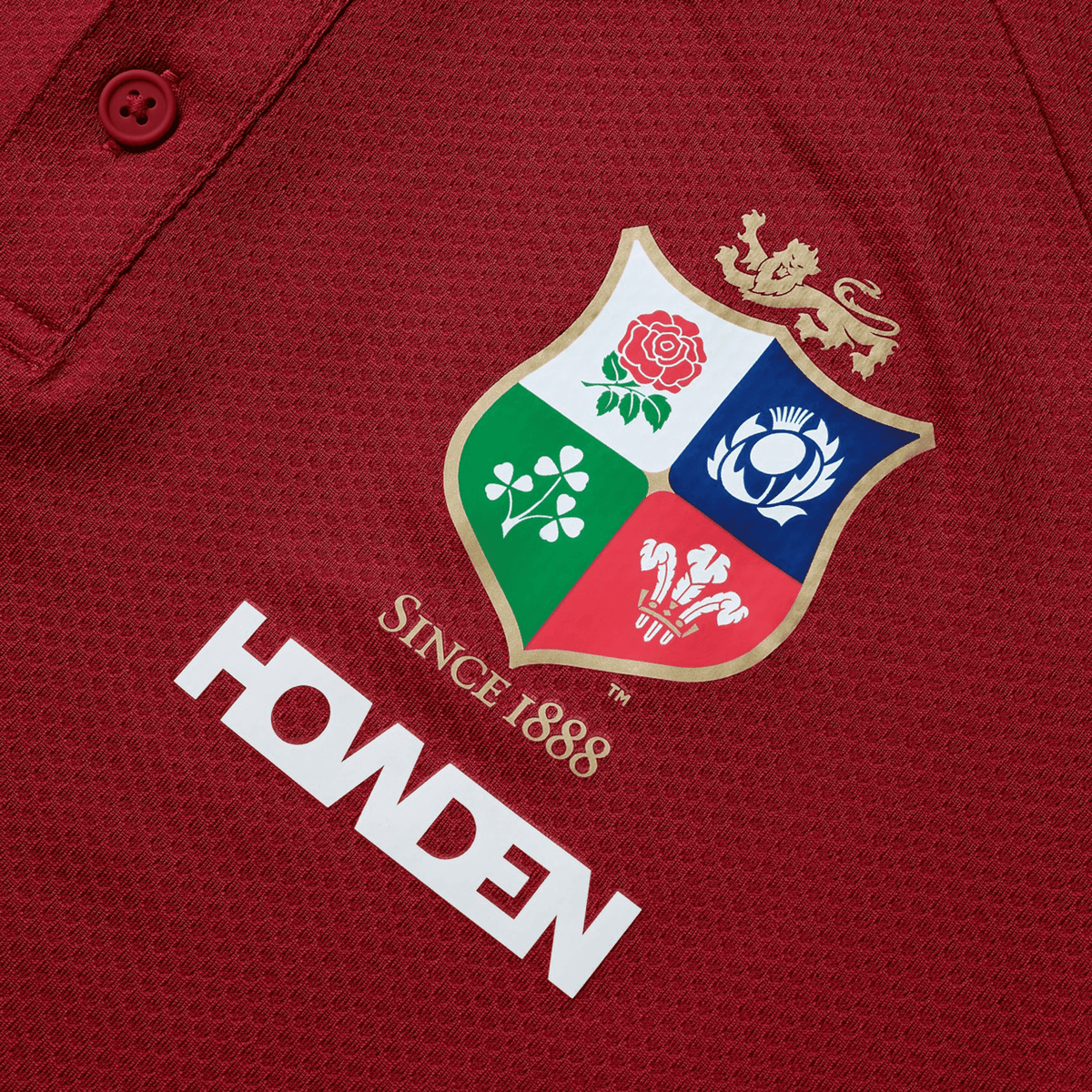 Close-up of a red shirt in breathable wicking fabric, featuring an embroidered shield with a lion, rose, clover, and thistle. Dated "Since 1888," with "HOWDEN" printed below, this design echoes the classic style of the British &amp; Irish Lions 25 Poly Polo by Canterbury.