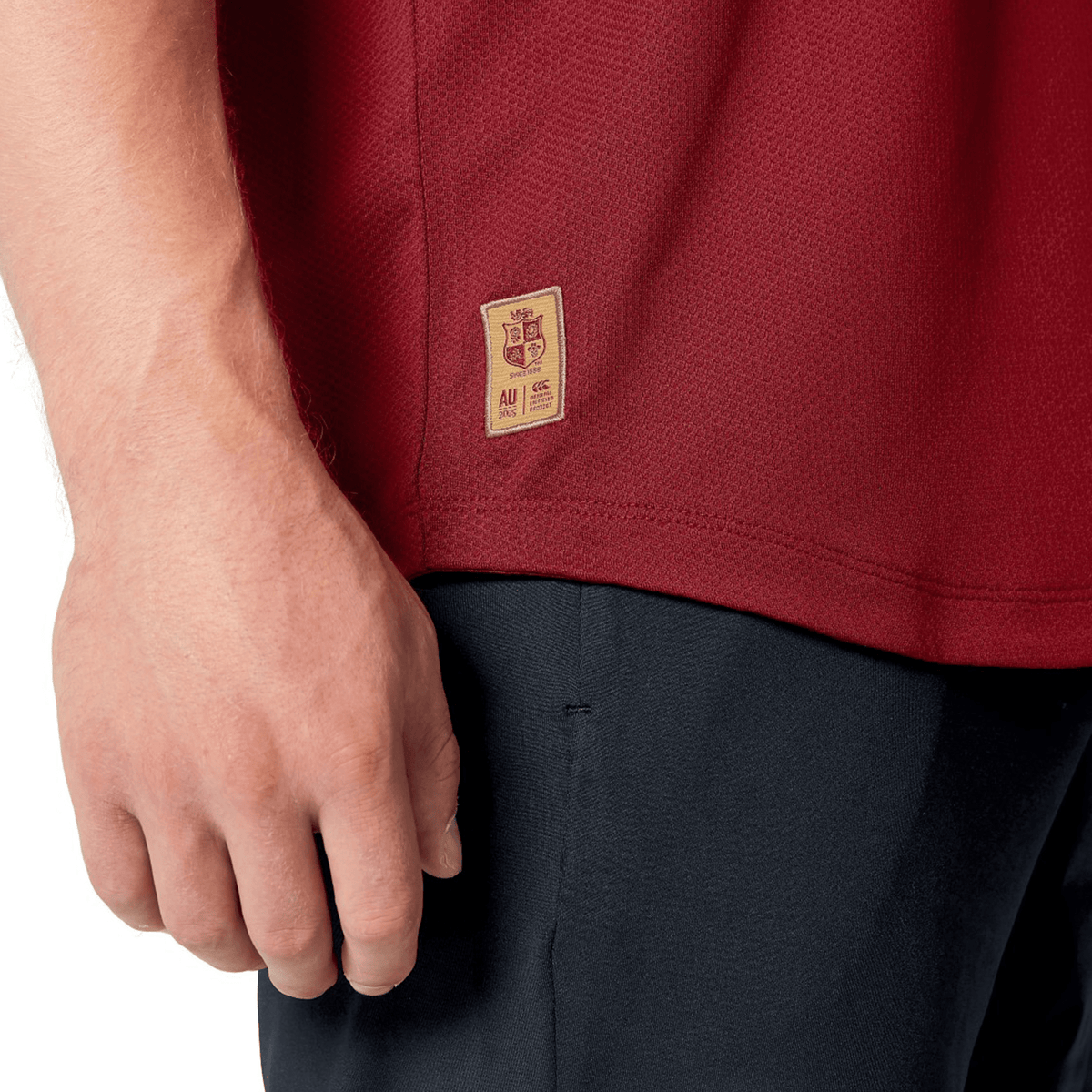 Close-up of a person’s hand by their side, wearing the red British &amp; Irish Lions 25 Poly Polo from Canterbury, featuring breathable fabric and a small tag, paired with black pants.
