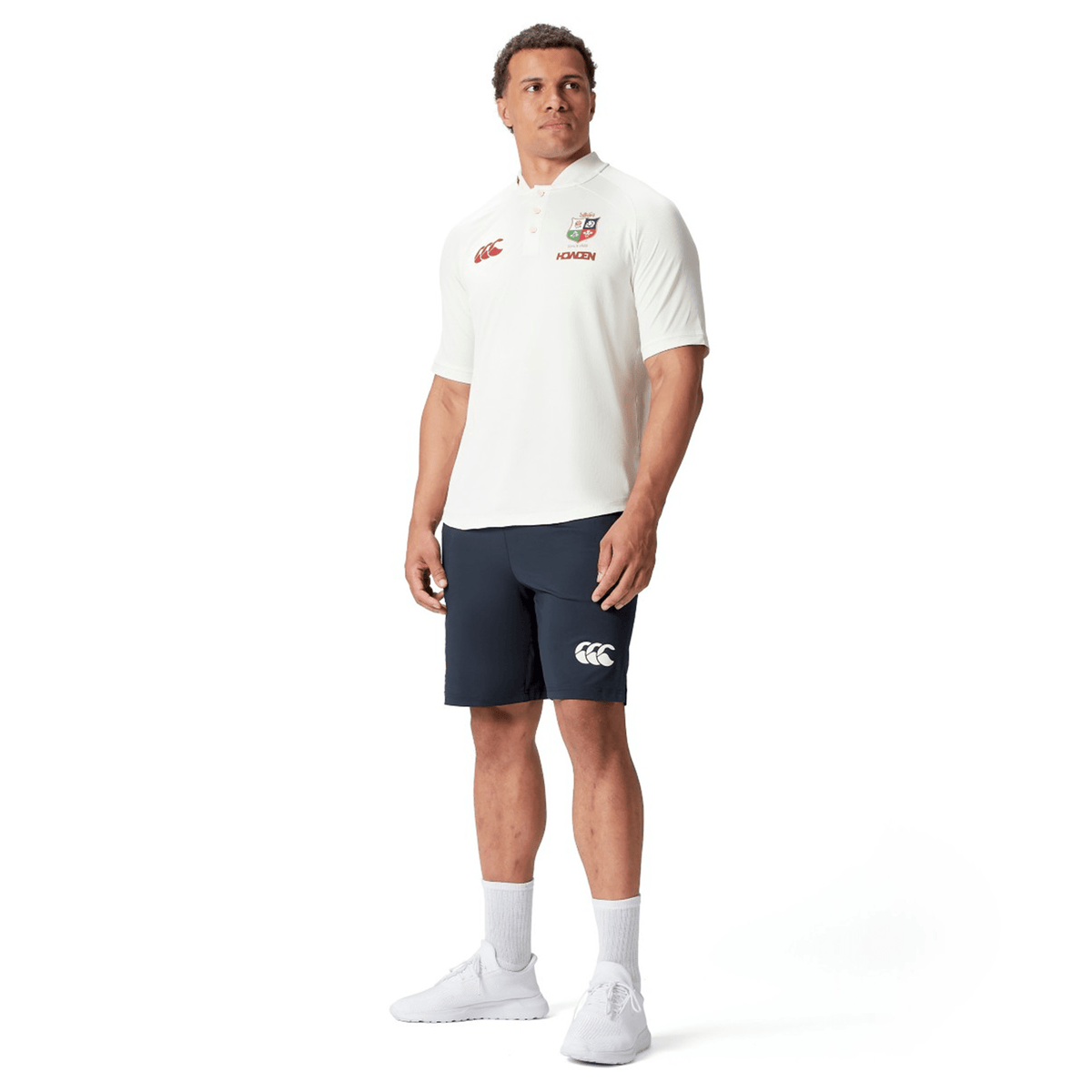 A man in sports attire stands confidently, wearing the British &amp; Irish Lions 25 Poly Polo by Canterbury, paired with navy shorts, white socks, and sleek white sneakers.