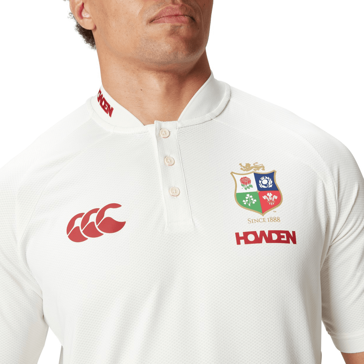 An individual is dressed in the British &amp; Irish Lions 25 Poly Polo by Canterbury, showcasing a white rugby shirt with a red Canterbury logo, the British and Irish Lions crest, and "Howden" branding. Its quick-drying fabric provides comfort during any match.