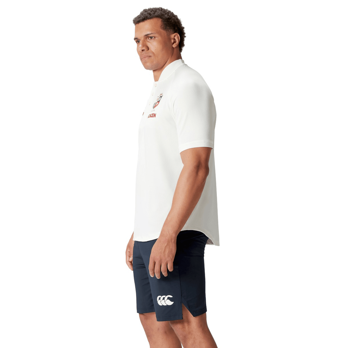 A man wearing a white British &amp; Irish Lions 25 Poly Polo by Canterbury and navy shorts stands against a white background, highlighting the embossed BIL neck tape.