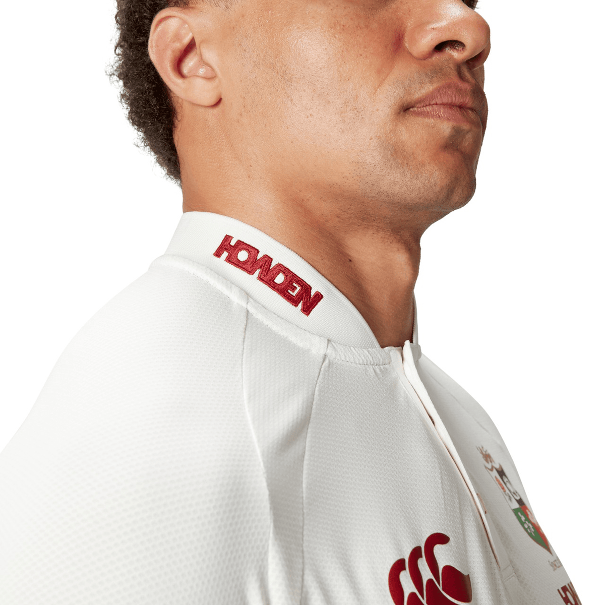 A side profile of a person wearing the British &amp; Irish Lions 25 Poly Polo by Canterbury, a white sports jersey adorned with red logos and text on the collar. Made with quick-drying fabric for enhanced comfort, this design features an embossed BIL neck tape that adds a refined touch.