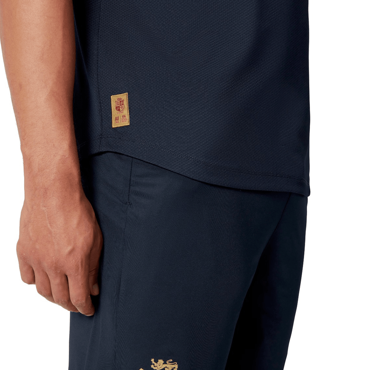 A close-up shot shows someone wearing the British &amp; Irish Lions 25 Poly Polo by Canterbury in a dark blue color with gold details. The quick-drying fabric of the polo is highlighted, along with their left hand positioned near the side seam logo on the shirt, showcasing Canterbury's commitment to quality.