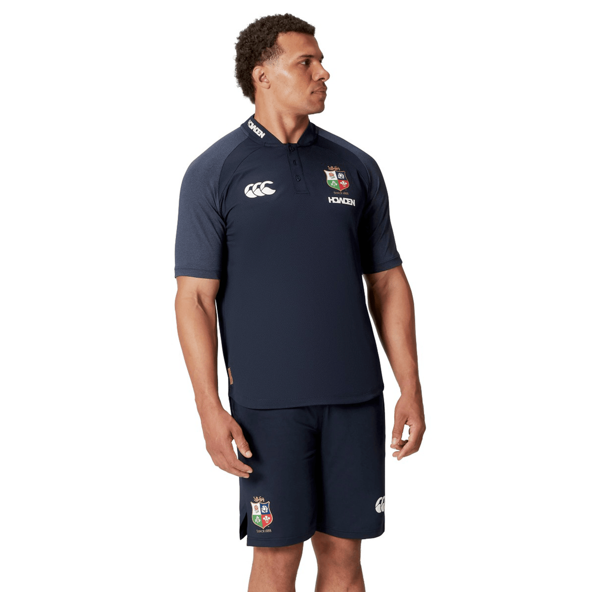 A man wearing the British &amp; Irish Lions 25 Poly Polo, a navy blue sports shirt featuring the Canterbury logo and designed with breathable wicking fabric, stands against a white background.