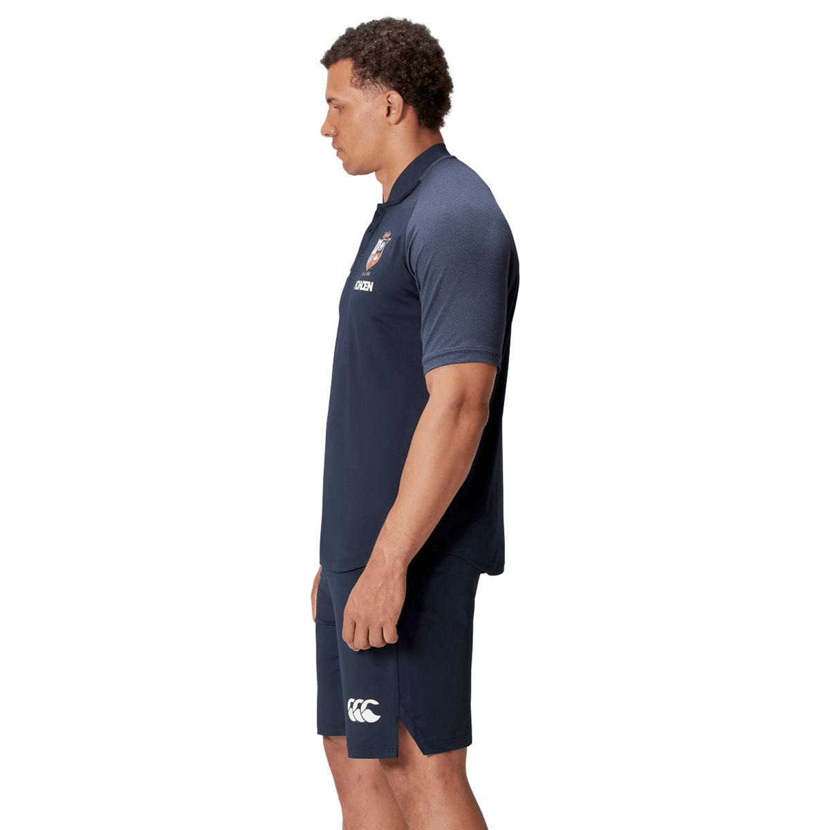 A man wearing the British &amp; Irish Lions 25 Poly Polo by Canterbury in navy, designed with breathable wicking fabric, stands sideways against a white background.