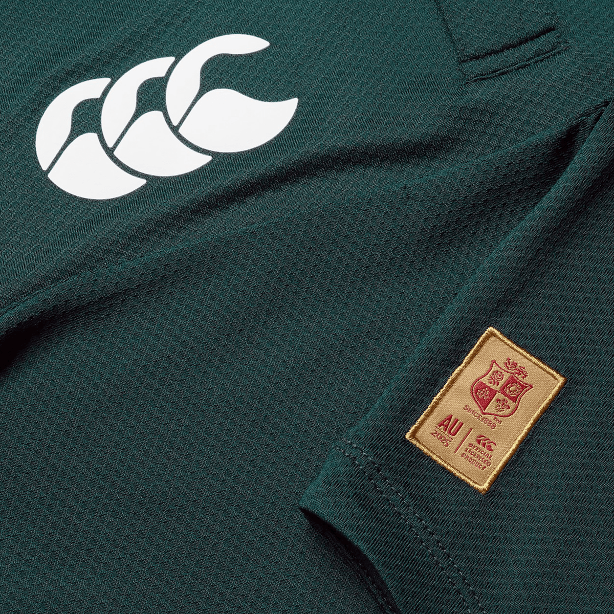 Close-up of a green sports jersey, crafted by Canterbury, featuring a white logo with three overlapping outlines and a red and gold patch displaying a crest and text. This British &amp; Irish Lions 25 Poly Polo is made from quick-drying fabric.
