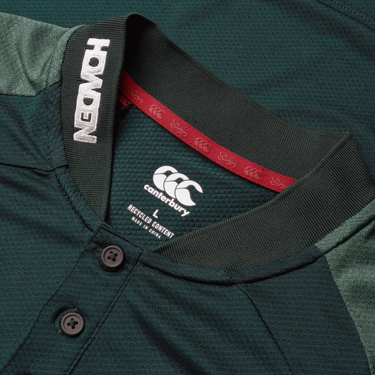Close-up of a green British &amp; Irish Lions 25 Poly Polo by Canterbury with black buttons, featuring "Howden" on the collar and a "recycled content" label inside. This quick-drying polo is ideal for those who value both performance and sustainability.