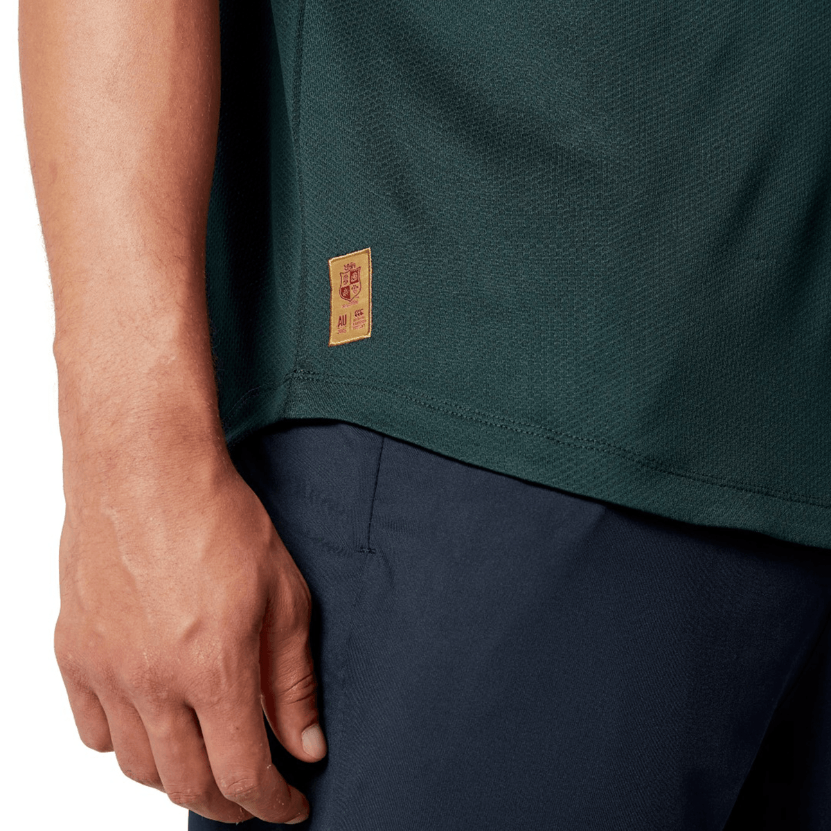 Close-up of a person wearing the British &amp; Irish Lions 25 Poly Polo by Canterbury, featuring a quick-drying dark green fabric and complemented by navy blue pants. A small patch is visible on the shirt as the individual's hand rests by their side.