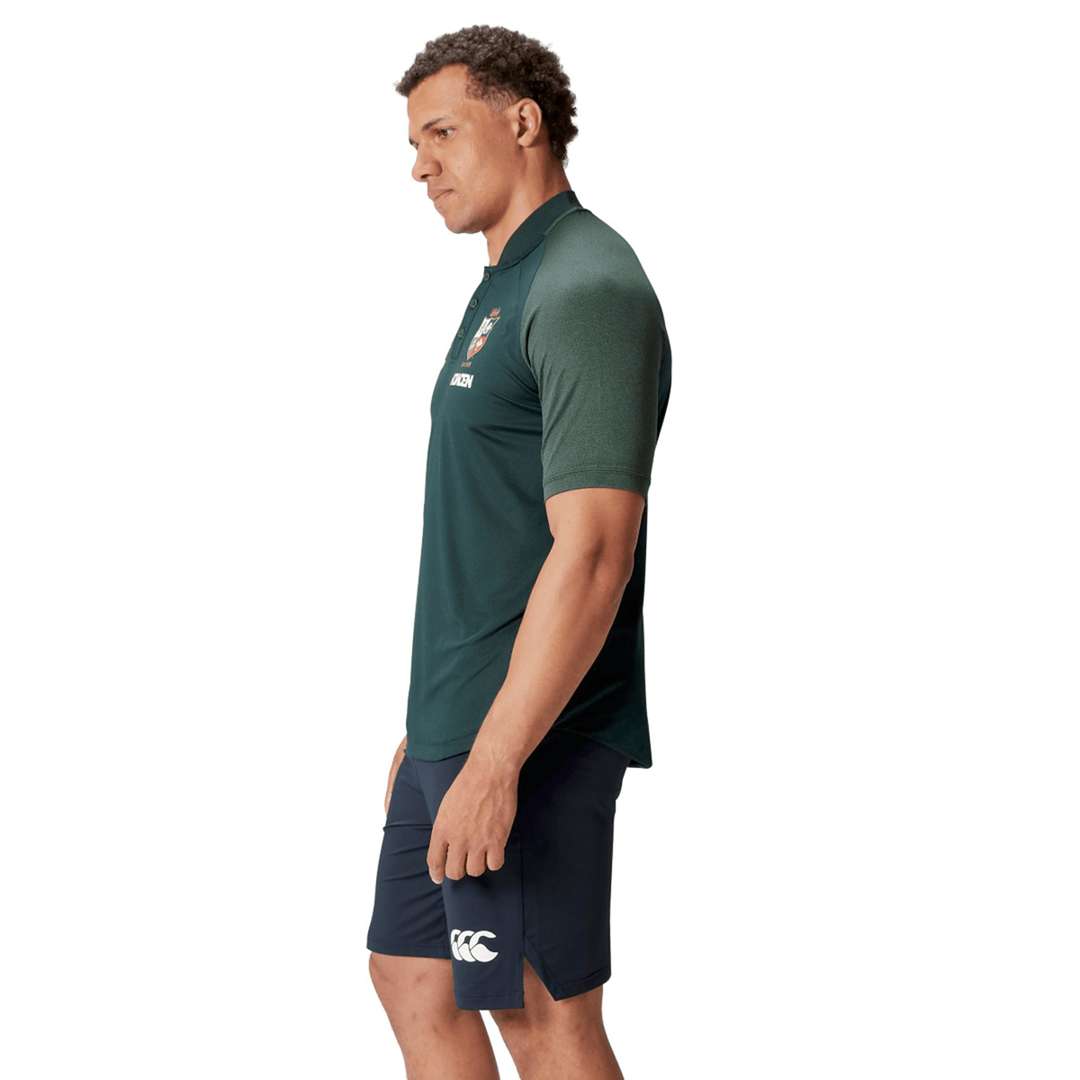 A man wearing the British &amp; Irish Lions 25 Poly Polo by Canterbury, a green collared sports shirt crafted from breathable wicking fabric, and navy shorts stands in a side profile against a white background.