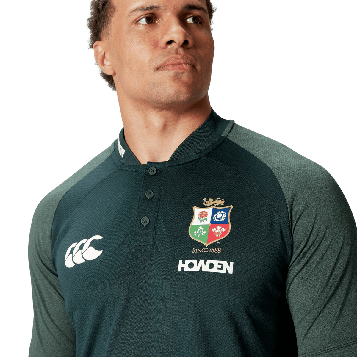 A person wearing the British &amp; Irish Lions 25 Poly Polo by Canterbury, featuring a dark green color with a multicolored crest and brand logos on the front. Made from breathable, wicking fabric, it promises maximum comfort and quick drying while keeping your style effortless.