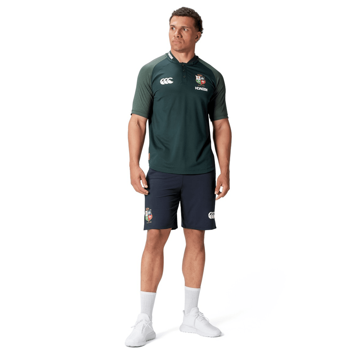 A man stands wearing the British &amp; Irish Lions 25 Poly Polo by Canterbury, a dark green and black sports uniform made of breathable wicking fabric, adorned with logos. He pairs it with white socks and sneakers against a white background.