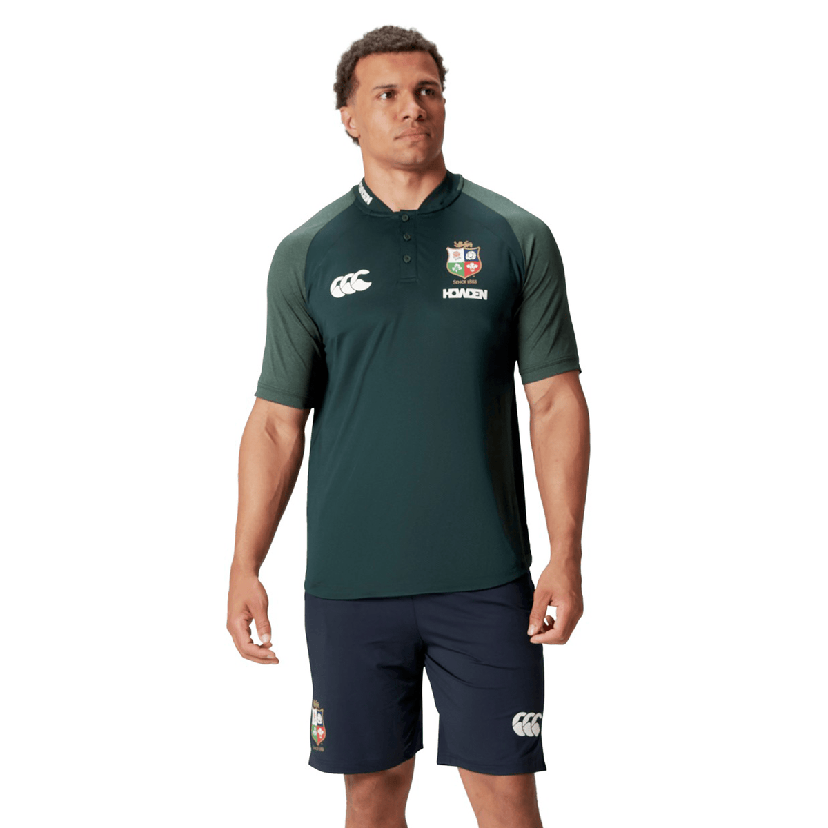 A person wearing the British &amp; Irish Lions 25 Poly Polo by Canterbury, a green and black sports shirt featuring a logo and made from breathable wicking fabric, paired with navy shorts, stands against a white background.