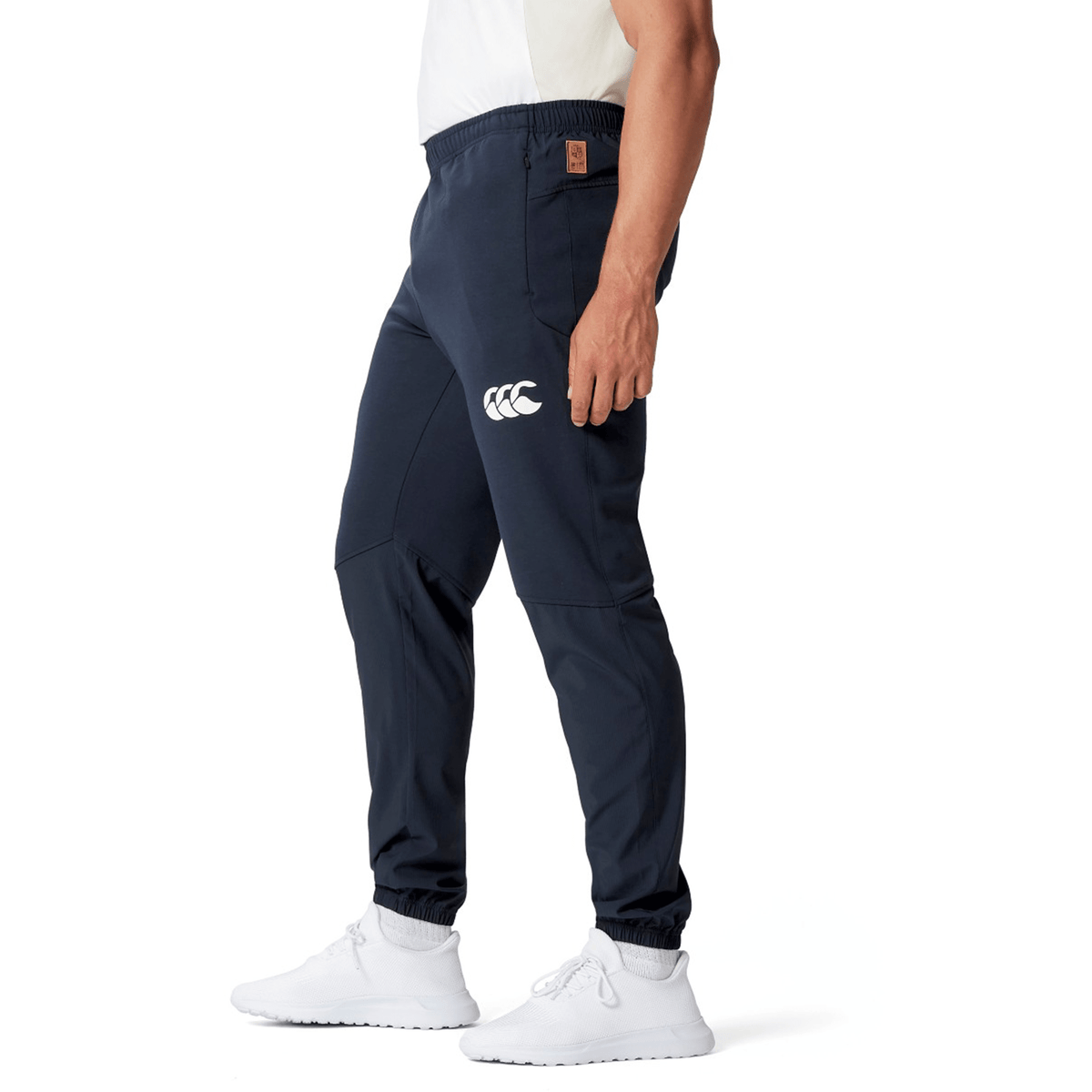 A person wearing the British &amp; Irish Lions 25 Training Pants by Canterbury in navy color, featuring a white Canterbury logo, paired with white sneakers, standing sideways.