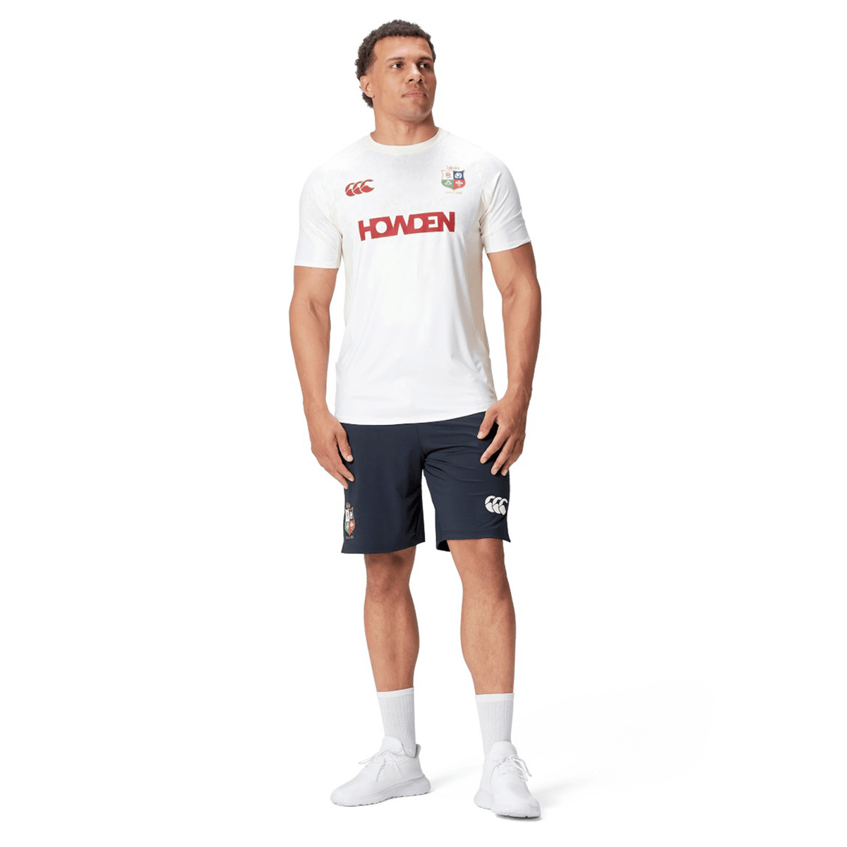 A man is wearing a white sports t-shirt with "Howden" printed in red and British &amp; Irish Lions 2025 Training Shorts by Canterbury, standing against a white background.