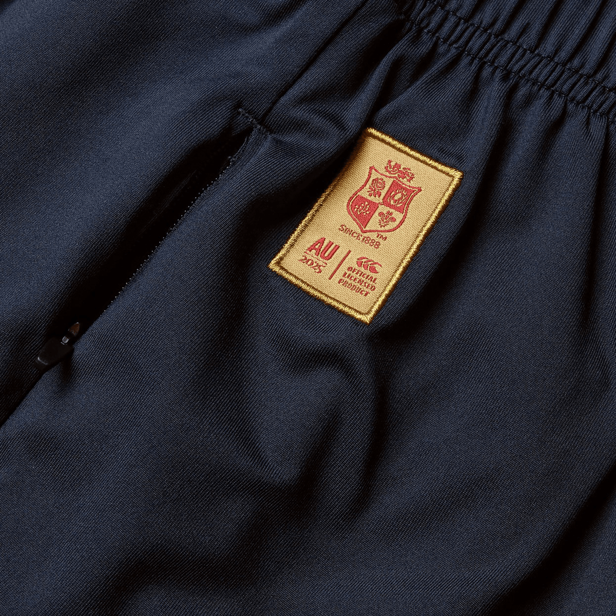 Close-up of navy blue pants by Canterbury, featuring an embroidered gold patch with a coat of arms and text details, reminiscent of the iconic British &amp; Irish Lions 2025 Training Short.
