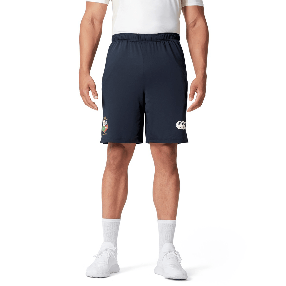 A person wearing a white shirt, British &amp; Irish Lions 2025 Training Short by Canterbury in a dark color with the logo visible, white socks, and white shoes stands against a white background.