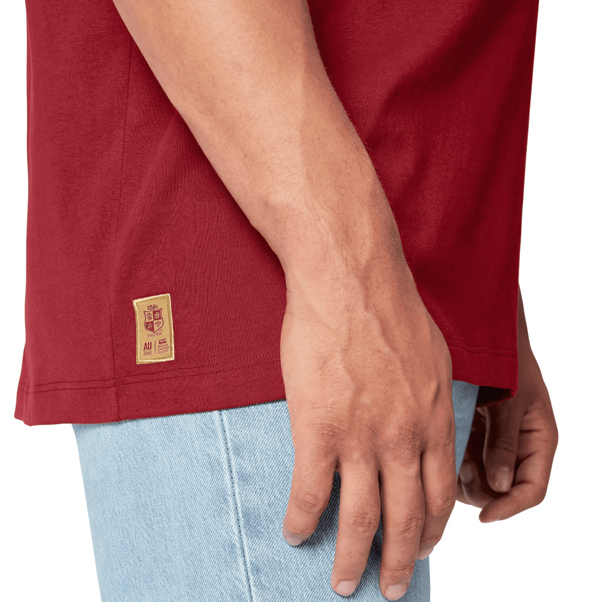 A person wearing a British &amp; Irish Lions 2025 Cotton Jersey Tee by Canterbury, made of organic cotton, in red along with light blue jeans. The tee resembles those worn by rugby supporters and features a small patch near the hem on the left side.